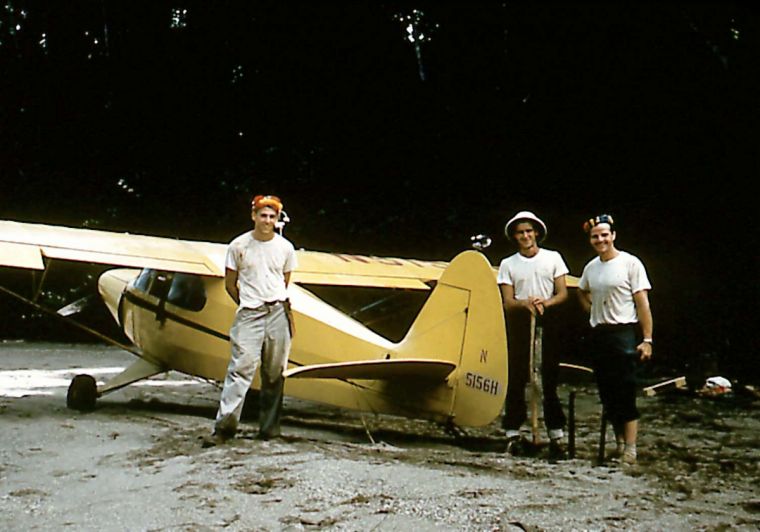 A vintage photograph showing three men standing beside a small yellow airplane on a dirt runway at night, with dark trees in the background.